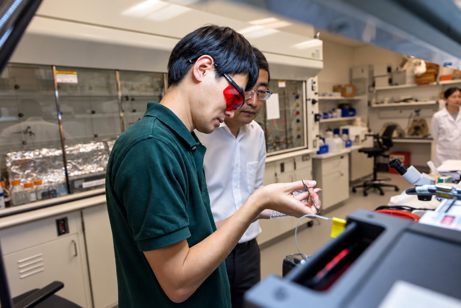 A researcher wearing red laser-safety goggles works at an optical rig while a colleague looks on